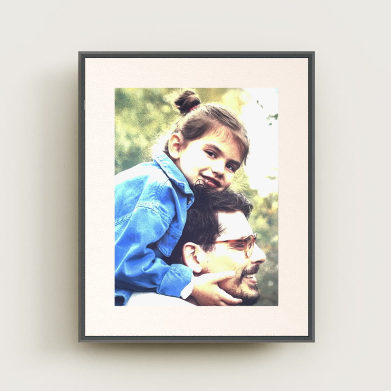 Aura Ink 13" cordless e-paper photo frame shown in portrait orientation, hung flat against a plain pale wall: a narrow graphite frame edge around a wide off-white paper-textured mat, with a color photograph of a small girl leaning over an adult's head filling the display; a soft shadow falls below and to the left and nothing else is in the shot