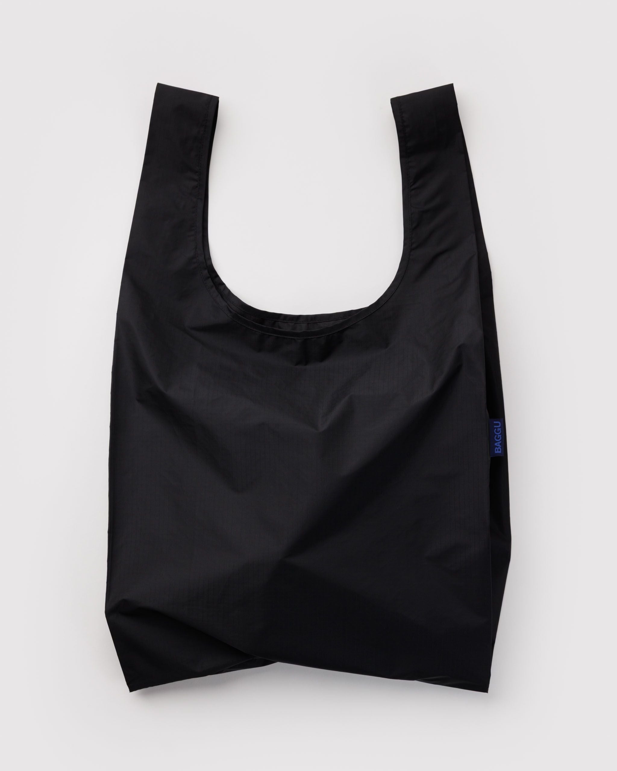 Black Standard Baggu reusable bag laid open on a pale gray backdrop, handles at the top and a small navy BAGGU tag at the right seam