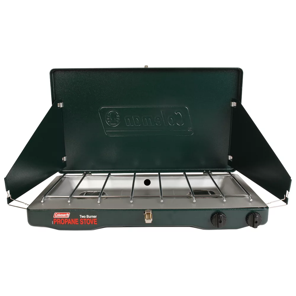 Green Coleman two-burner propane camp stove standing open on a white background, lid raised and side wind panels folded out over a chrome grate