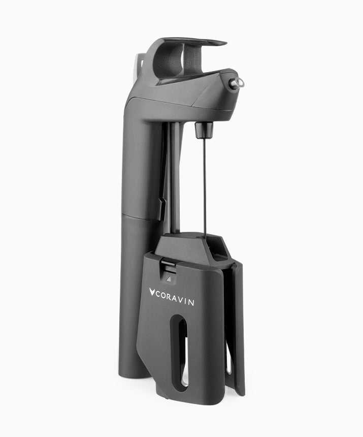 Matte black Coravin Timeless Three+ wine preservation system standing at a three-quarter angle on an off-white background, handle and trigger at the top, a thin needle pointing down into the clamp base printed CORAVIN