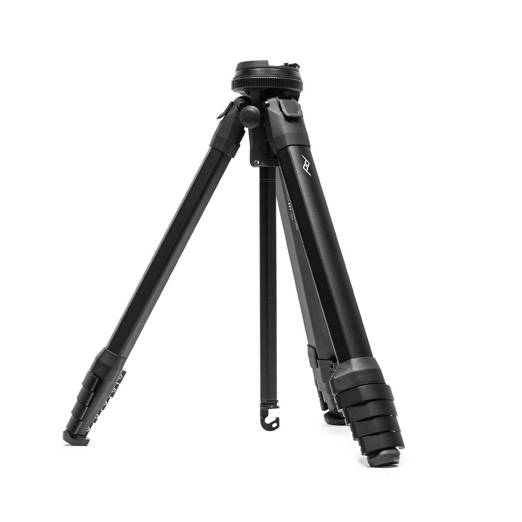 Peak Design Travel Tripod in black aluminum with its three legs opened, the compact ball head at the top, the Peak Design logo mark on one leg, and the load hook hanging below the center column, photographed against a white background
