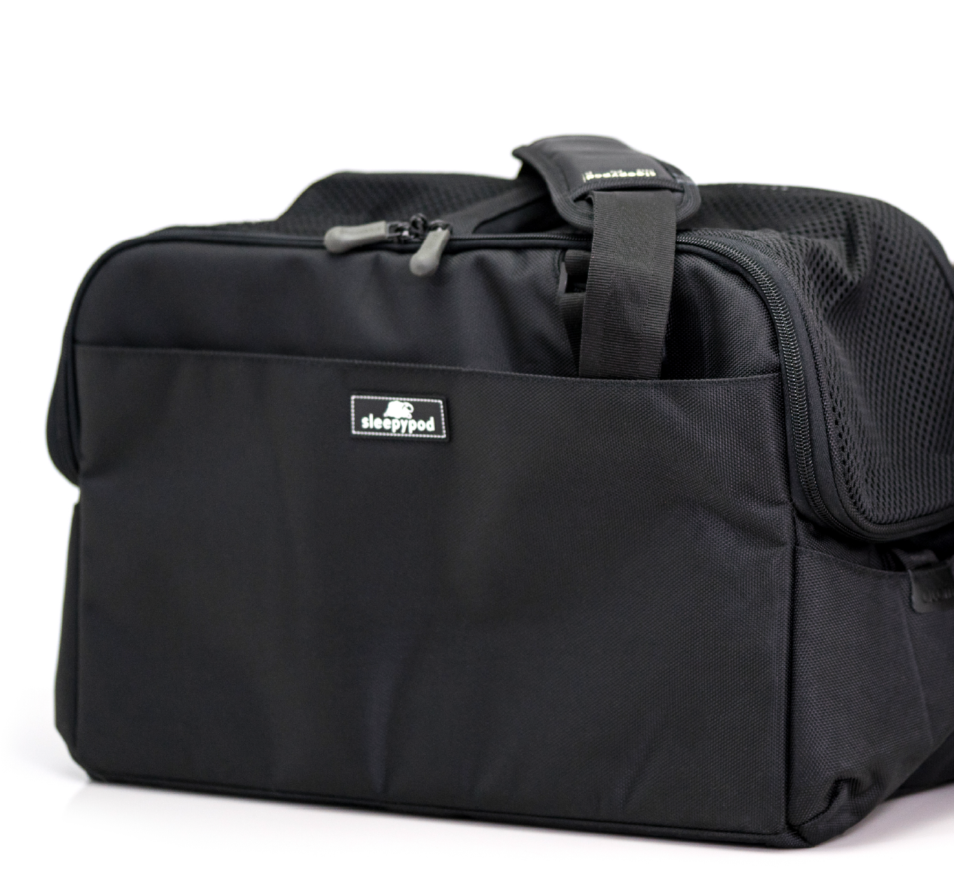 Sleepypod's Jet Black Atom carrier at a three-quarter front angle on a white background, cropped at the right edge — the padded top carry handle, two gray zipper pulls on the top zipper, the woven sleepypod label on the front pocket and part of the black mesh end panel